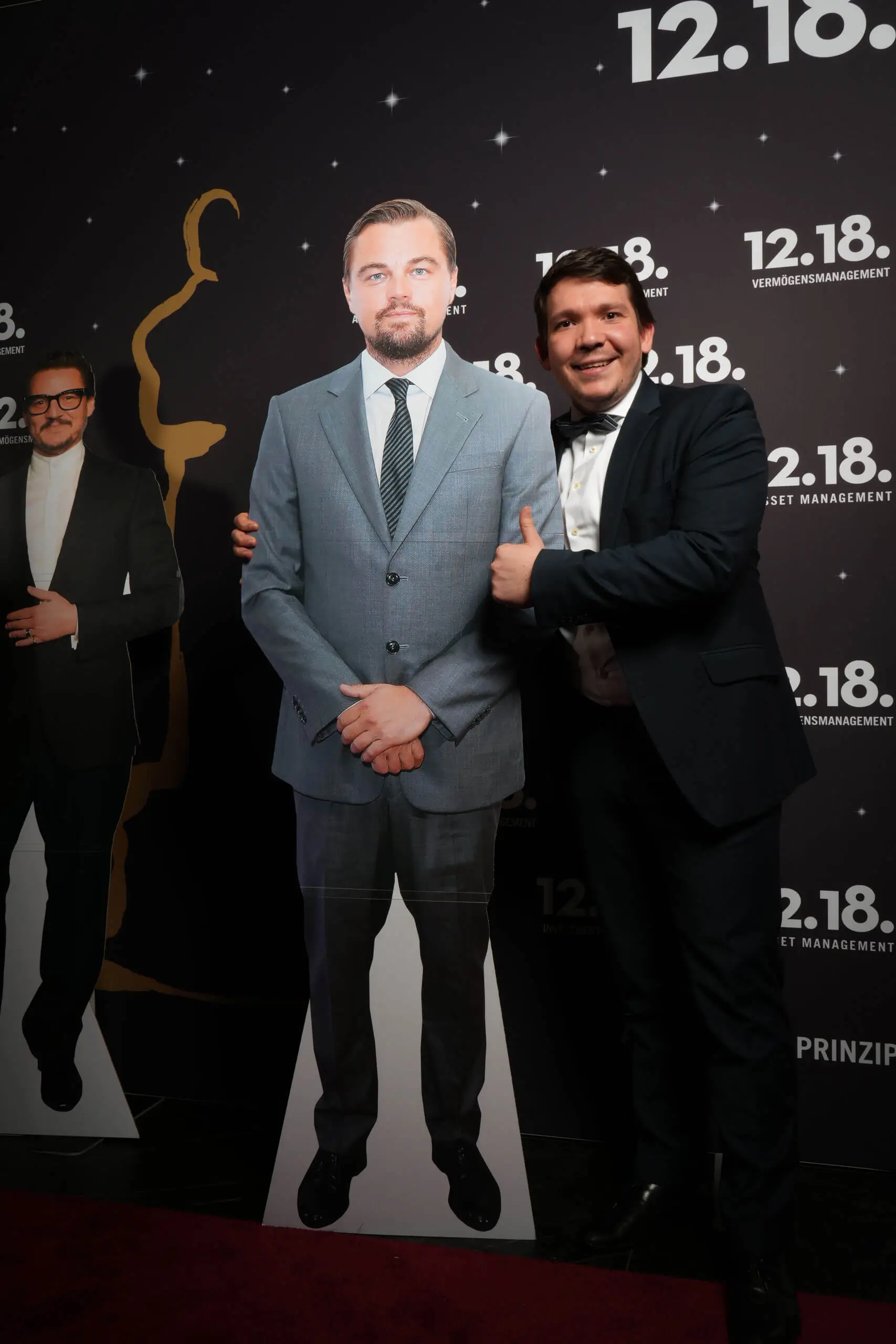 Mann posiert mit einer lebensgroßen Pappfigur von Leonardo DiCaprio bei einer eleganten Veranstaltung.
