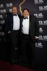 Two men standing in front of a wall with numbers, possibly related to investment management company 12.18. Investment, as indicated by the text on the image. The individuals are both wearing suits with ties, and one of them is smiling.