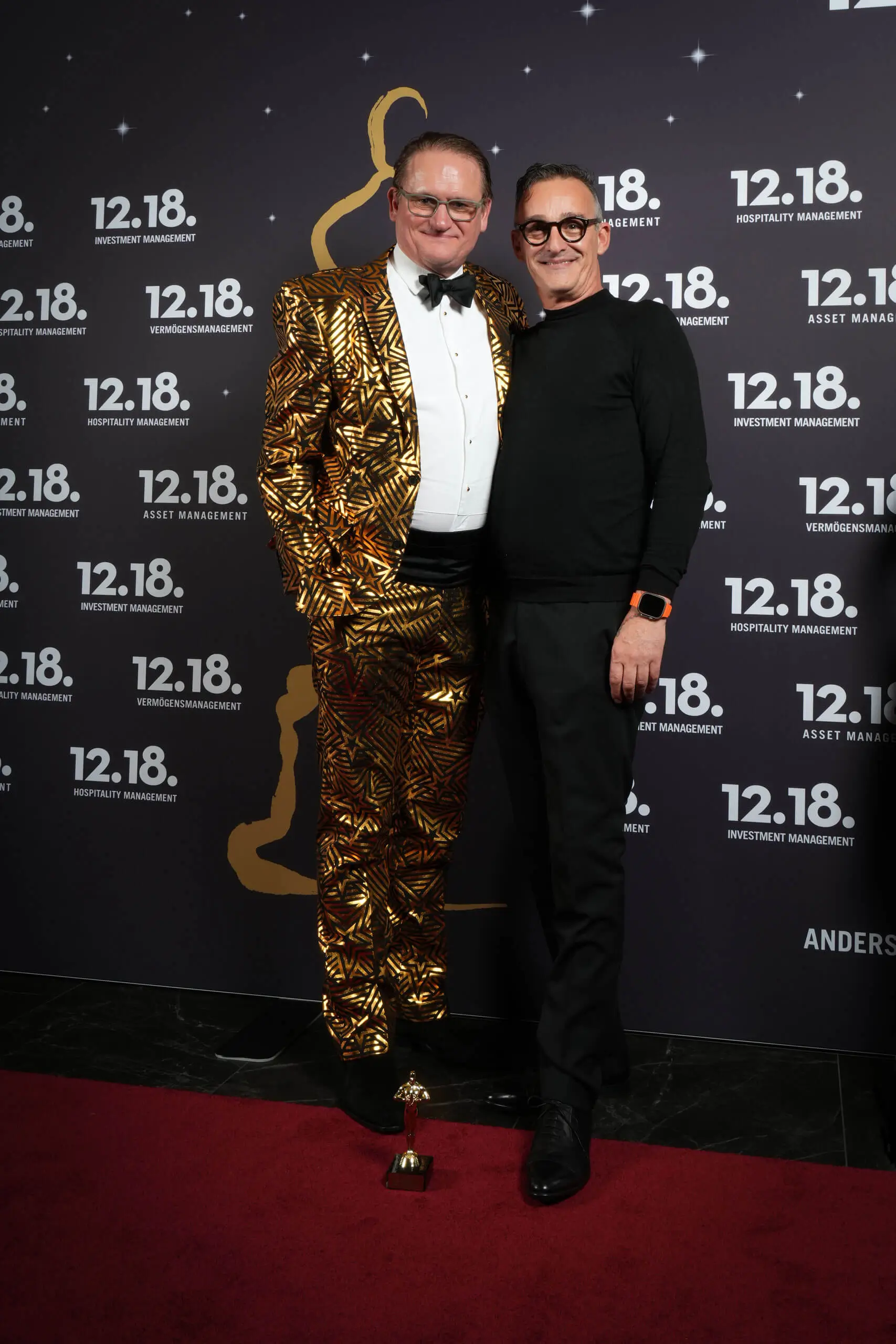 Zwei Männer in formeller Kleidung, einer in einem goldenen Anzug, posieren bei einer glamourösen Veranstaltung auf einem roten Teppich.