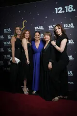 A group of women posing for a photo, possibly at a premiere event, wearing fashionable cocktail dresses and smiling. Context linked to 12.18. Investment Management GmbH.