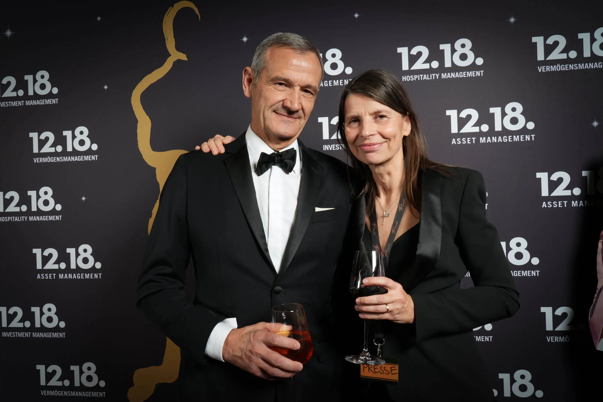 Elegantes Paar feiert bei einer Veranstaltung mit Drinks vor einem Branding-Hintergrund.
