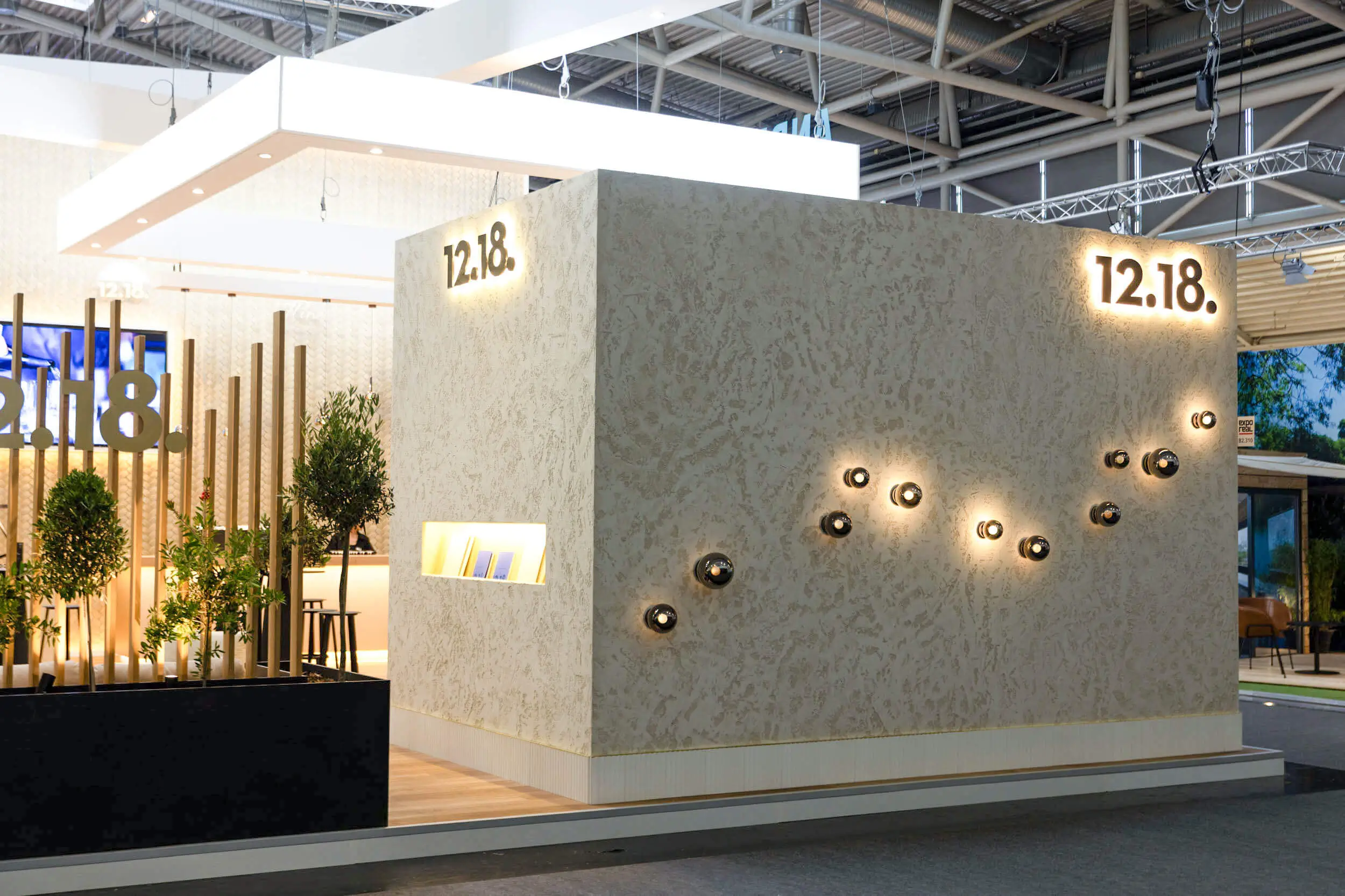Messestand mit 12.18.-Branding, dekorativer Wandbeleuchtung und Pflanzen.