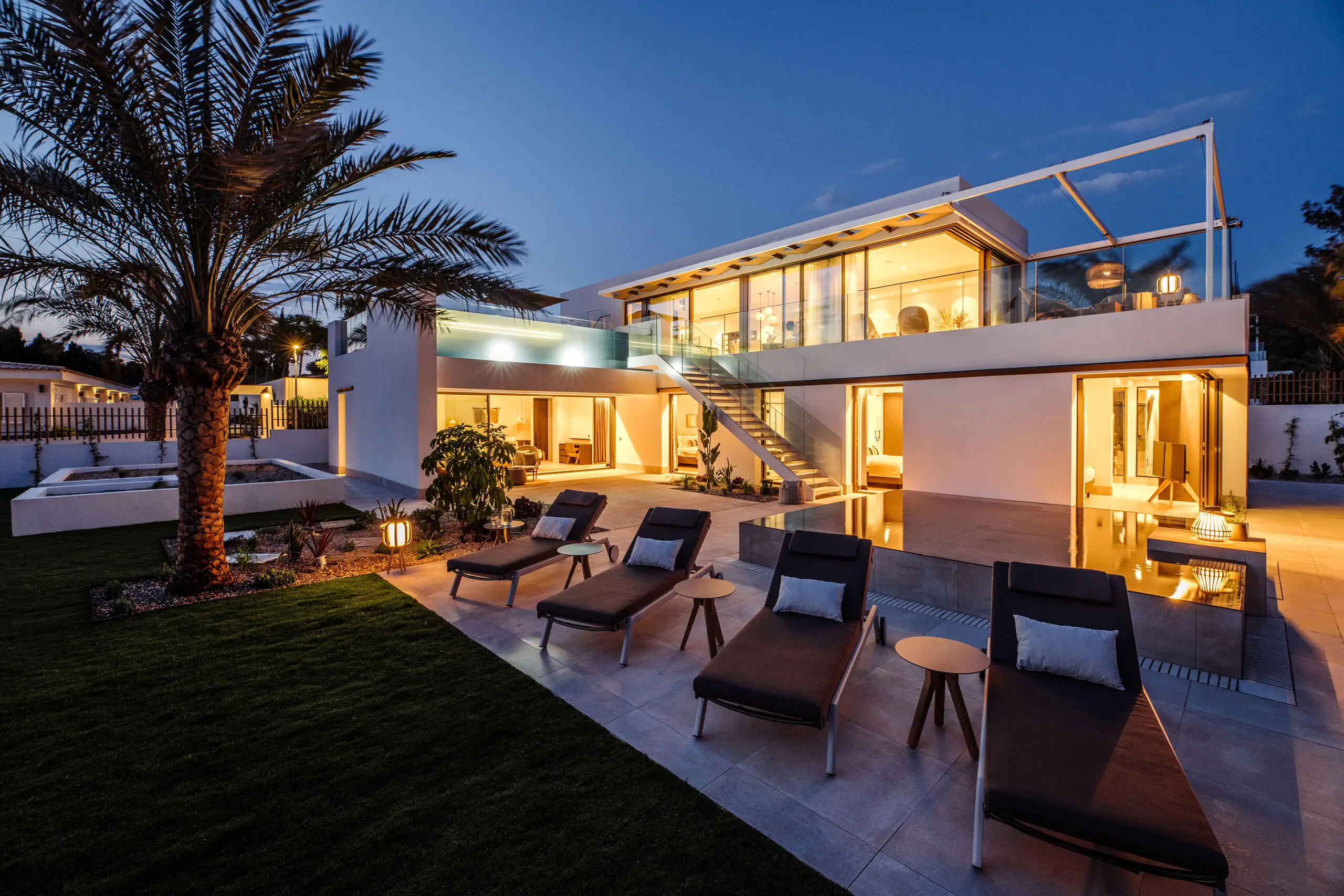Luxuriöse moderne Villa mit Palmen und Sonnenliegen am Pool in der Dämmerung.
