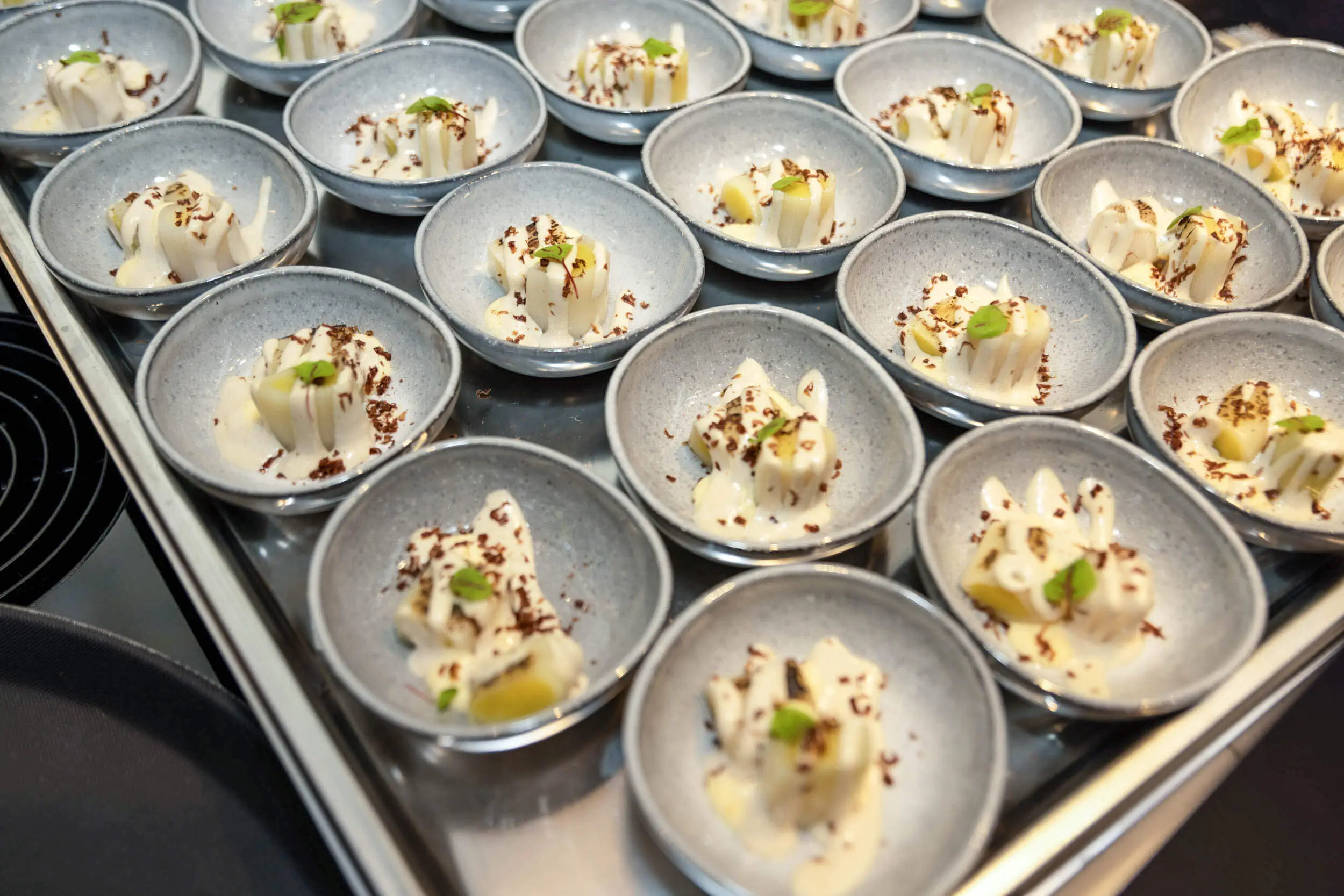 Viele kleine Dessertschalen mit cremigen Desserts, Schokoladenspänen und Kräutern auf einem Tablett.