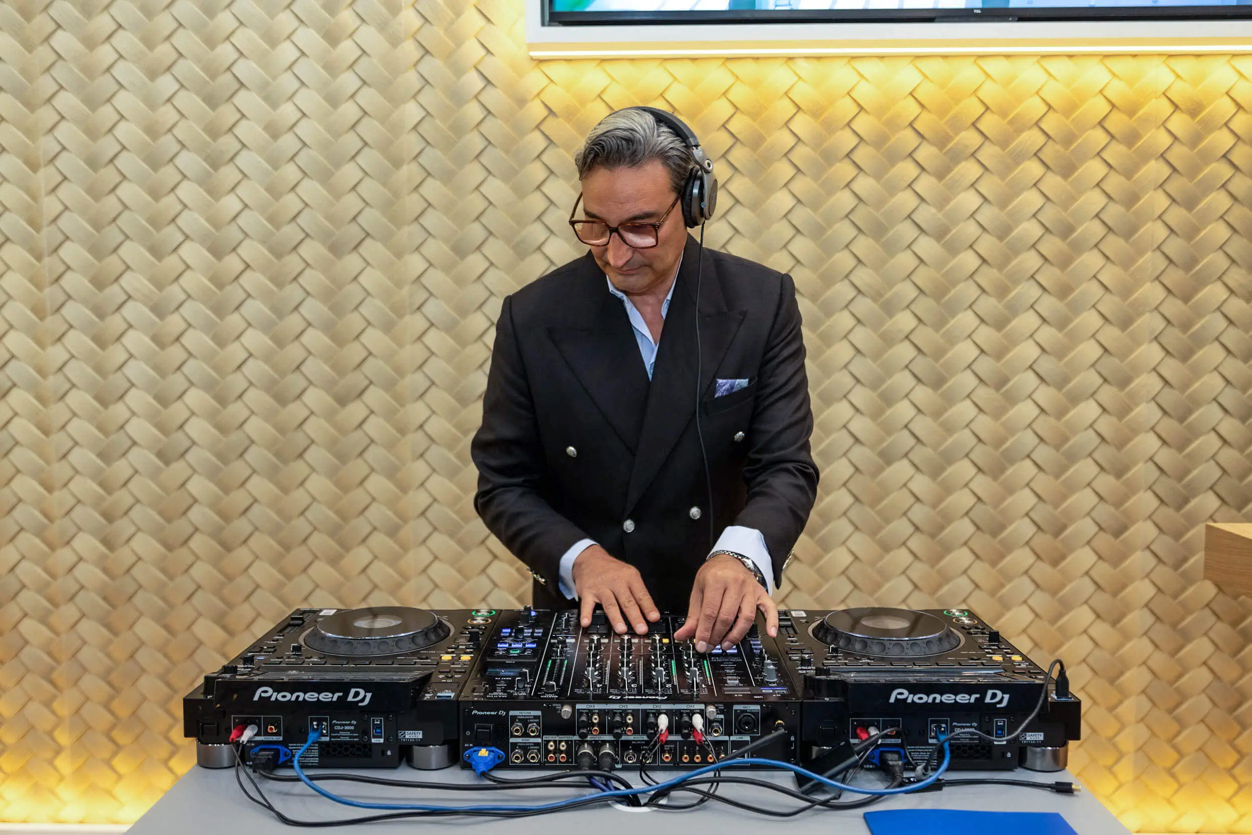 Ein DJ mit Kopfhörern bedient an einer Pioneer-DJ-Anlage ein Mischpult vor einer goldenen Wand.
