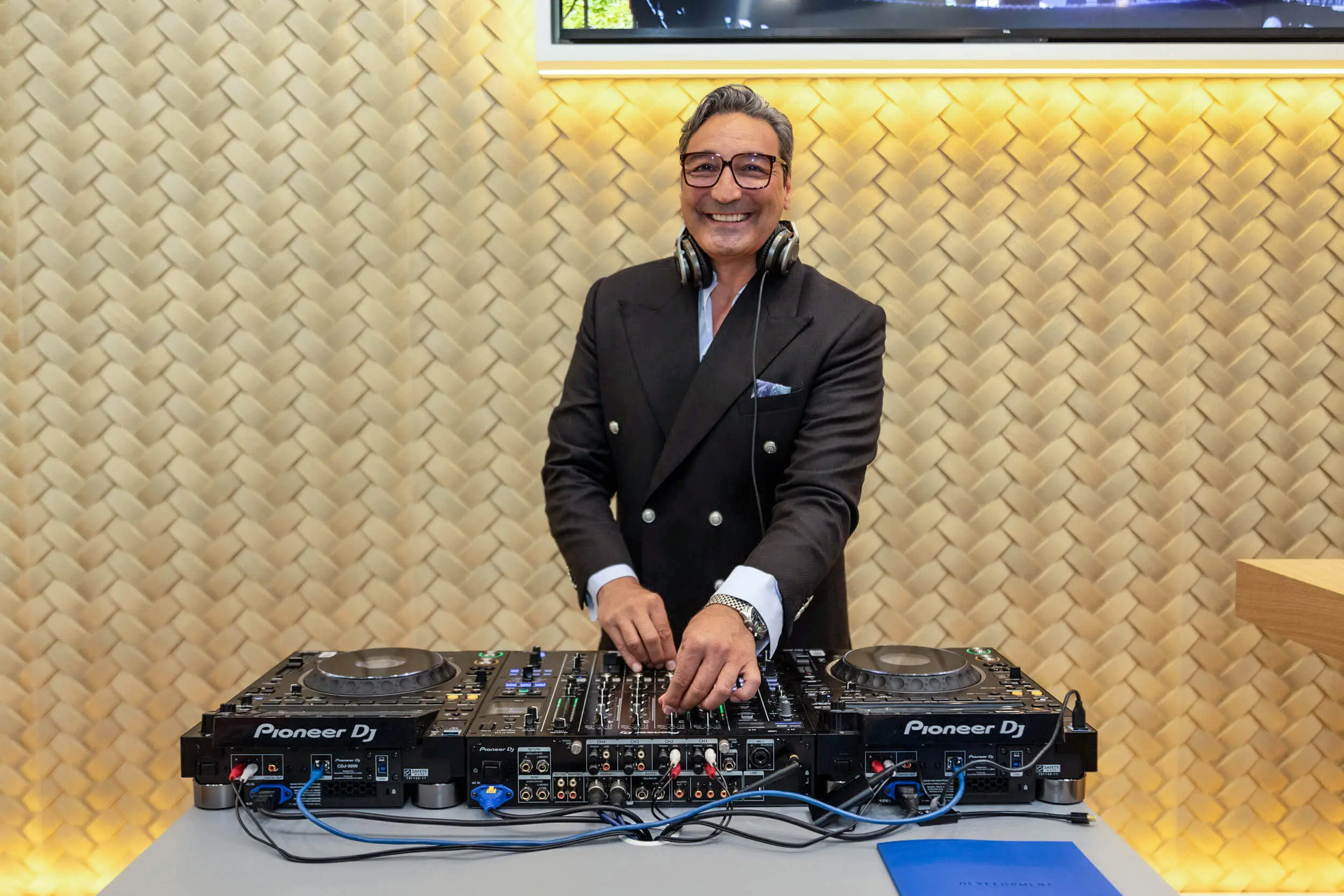 Ein lächelnder DJ im Anzug steht an einem Pioneer-DJ-Setup bei einer Veranstaltung.