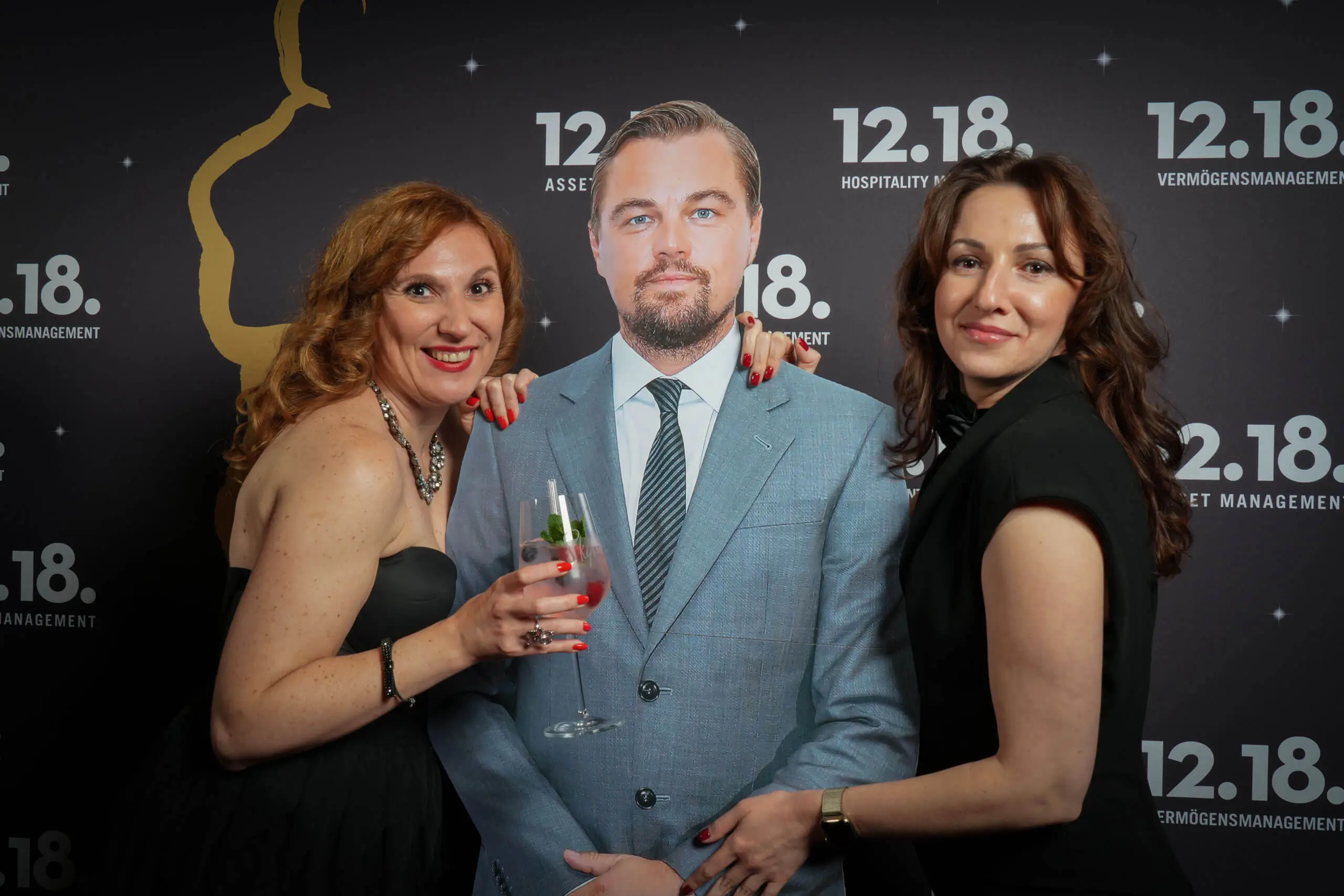 Zwei Frauen lächeln mit einem Pappaufsteller von Leonardo DiCaprio bei einer Veranstaltung.
