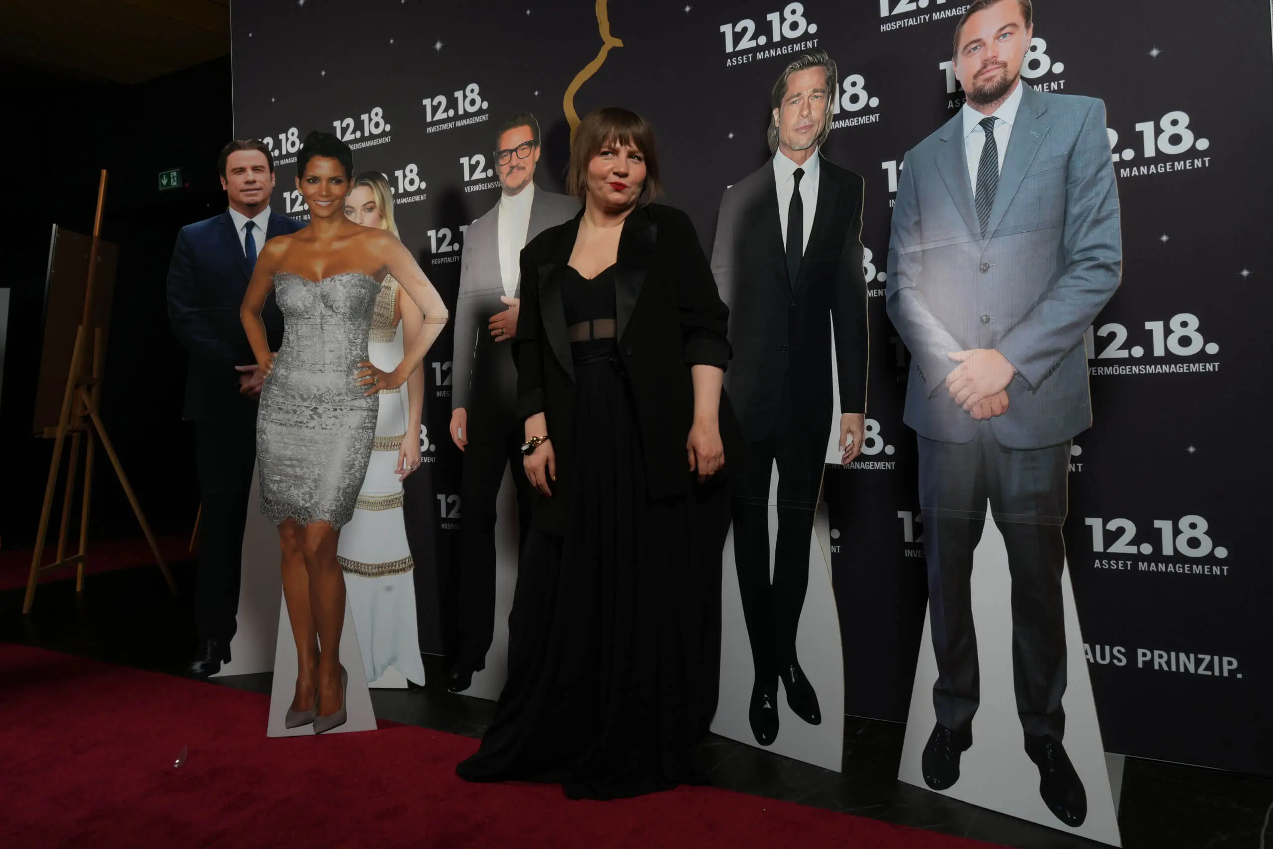 Eine Frau in einem schwarzen Kleid posiert mit Prominenten-Aufstellern bei einer Vermögensverwaltungsveranstaltung.