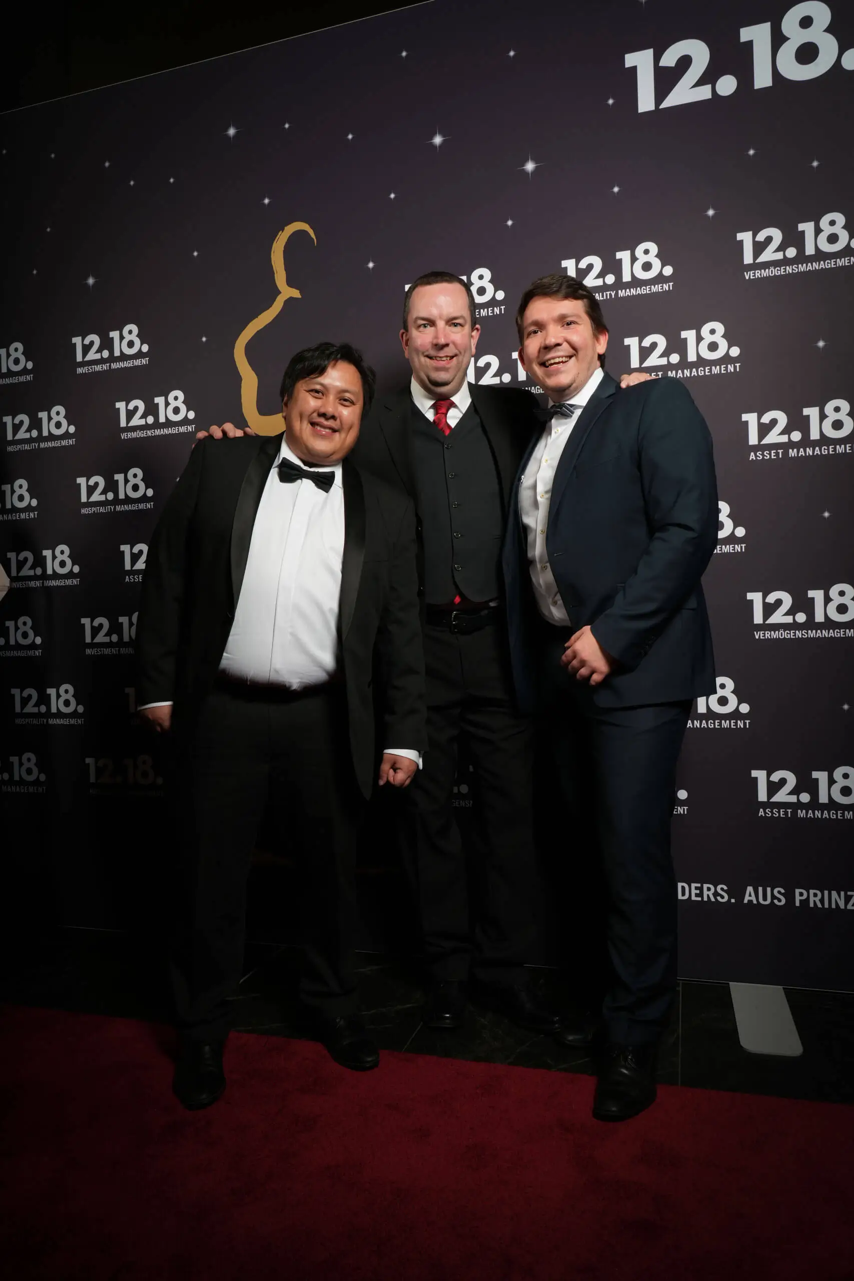 Drei Männer in formeller Kleidung posieren auf einer Gala-Veranstaltung und lächeln in die Kamera.