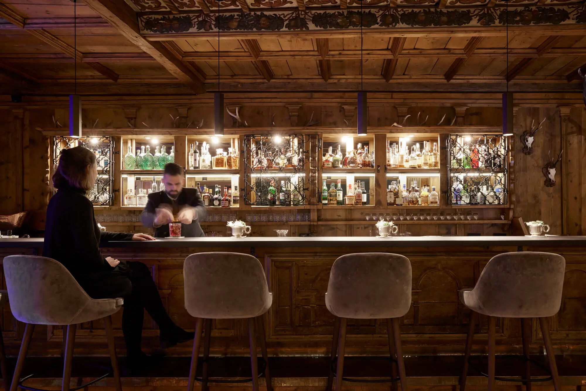 A bartender mixes drinks at a cozy bar with shelves of liquor bottles.