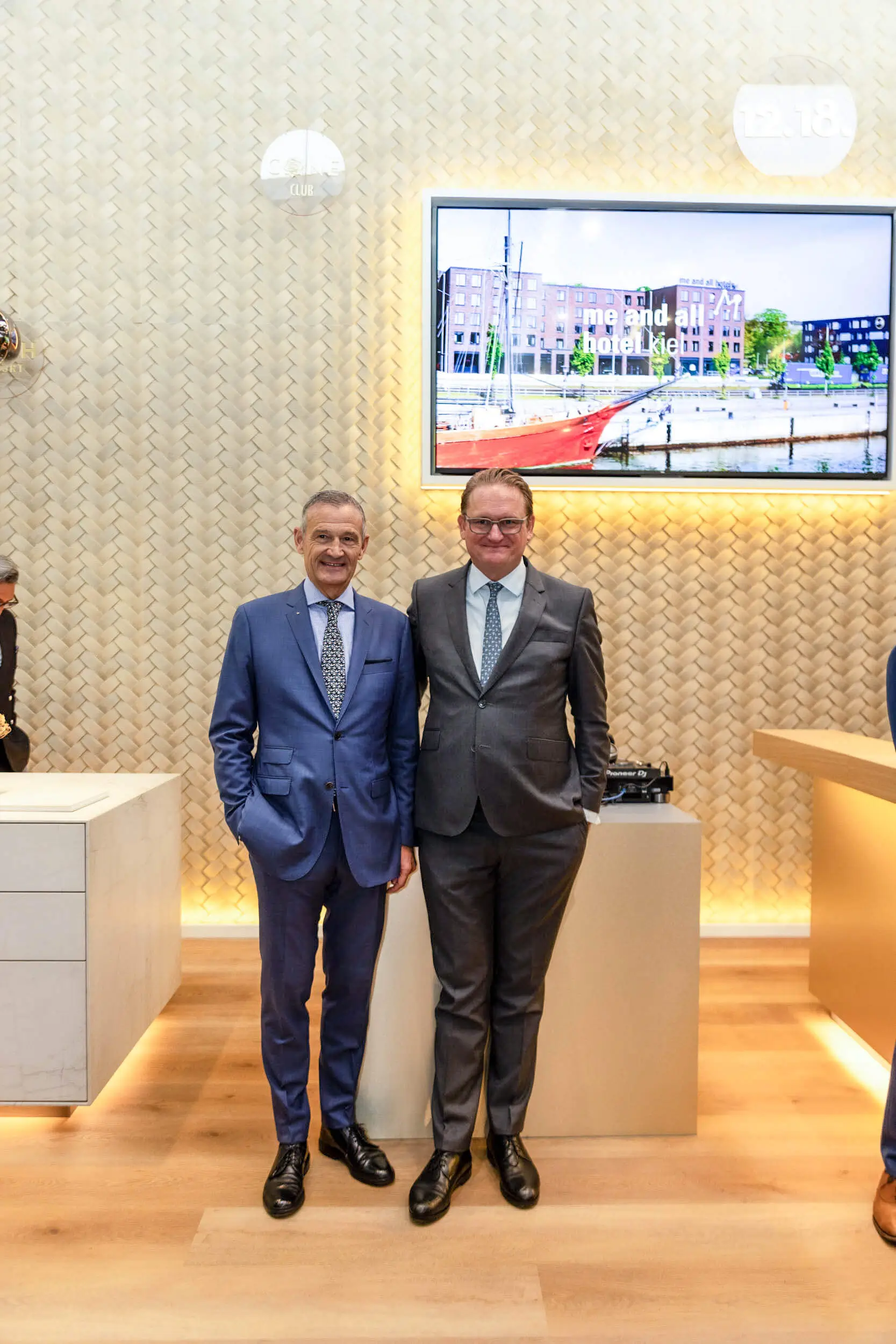 Zwei Geschäftsleute im Anzug stehen in einer hellen Hotellobby vor einem Wanddisplay.