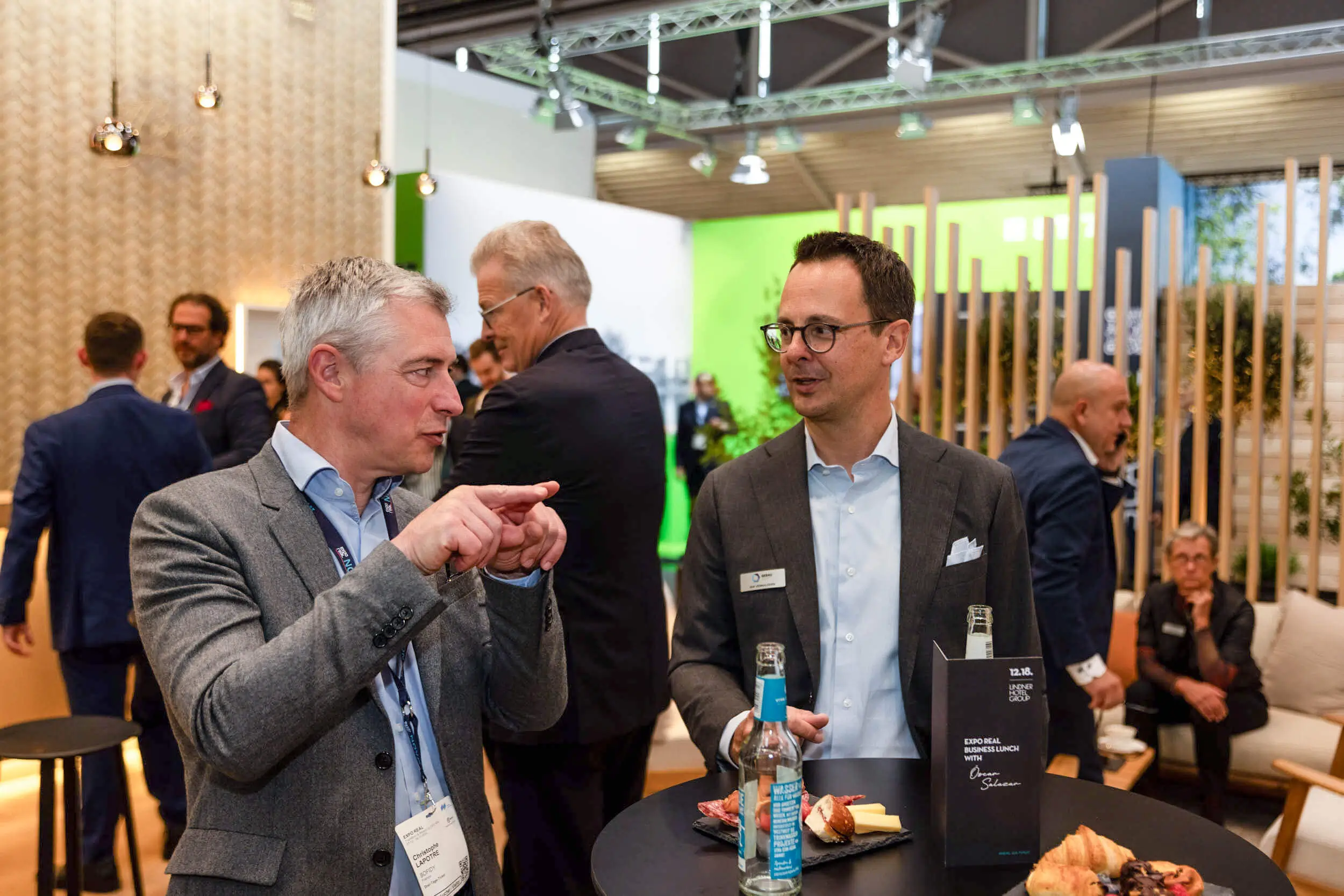 Zwei Männer im Anzug sprechen an einem Messestand mit Getränken und Snacks.