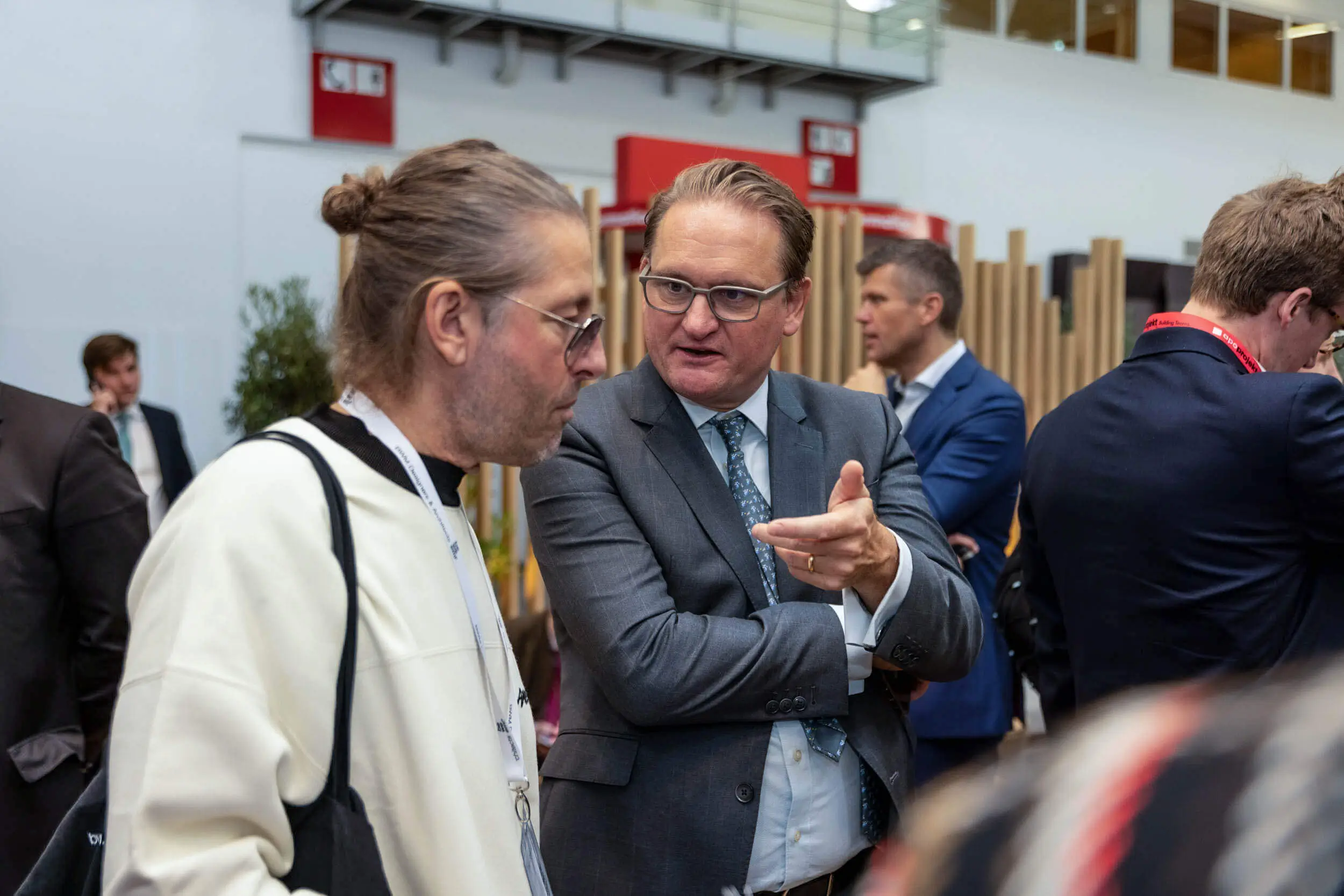 Zwei Männer sprechen auf einer Konferenz, im Hintergrund weitere Teilnehmer.