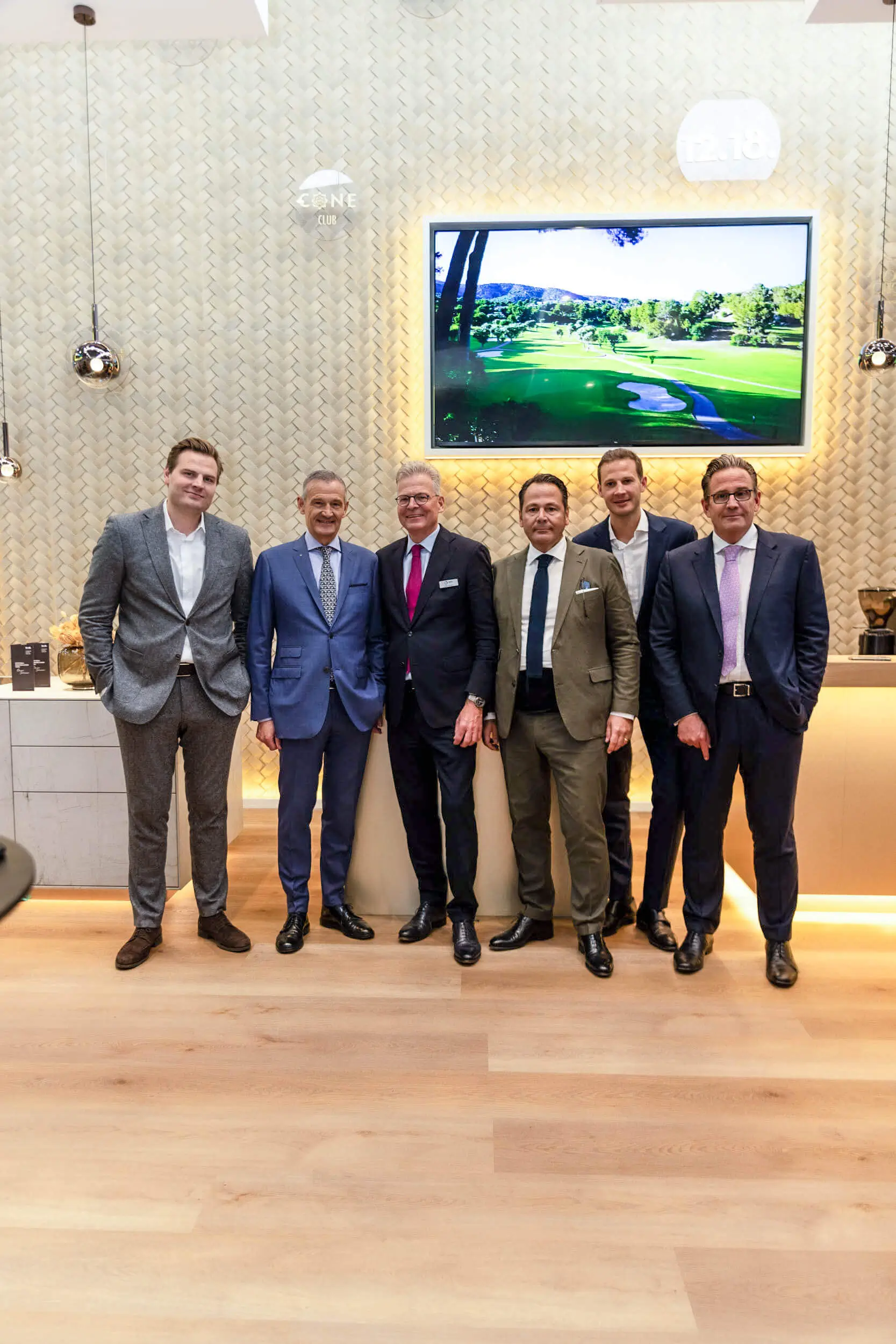 Sechs Männer in Anzügen posieren bei einem Business-Event vor einem Bildschirm mit Golfplatzmotiv.