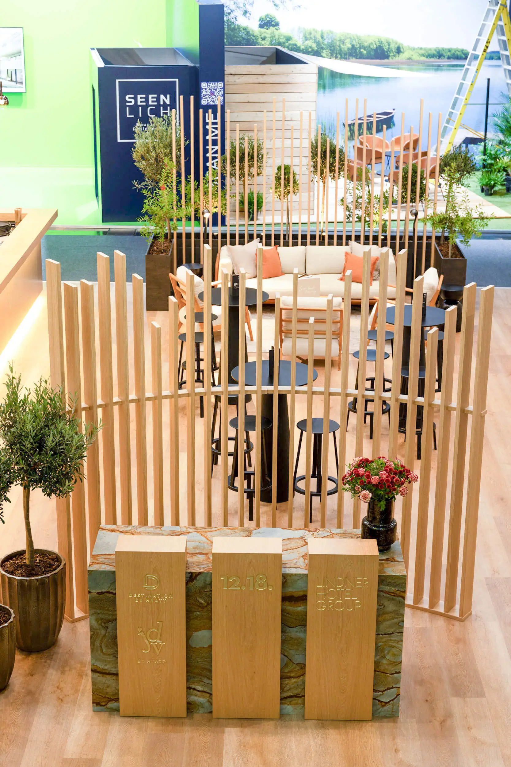 Offene Messelounge mit Holzlamellen, weißem Sofa, Stehtischen und dekorativen Pflanzen vor einer gebrandeten Expo-Wand.