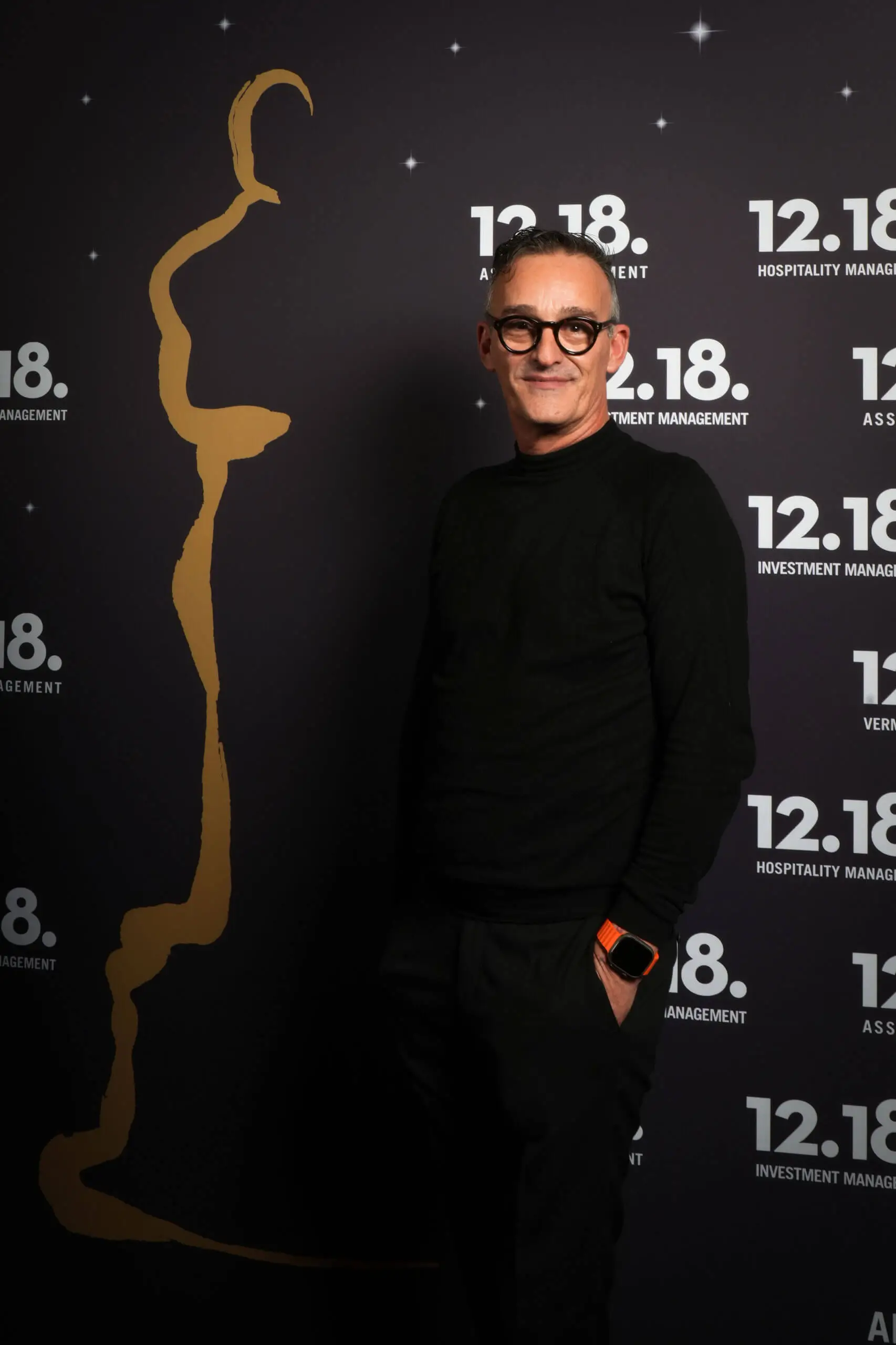 Ein Mann in einem schwarzen Pullover posiert vor einem eleganten Hintergrund für eine Veranstaltung.