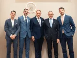 A group of well-dressed men standing in a semi-circle, wearing suits and ties. Image is linked to the 12. 18. Investment Management GmbH website.