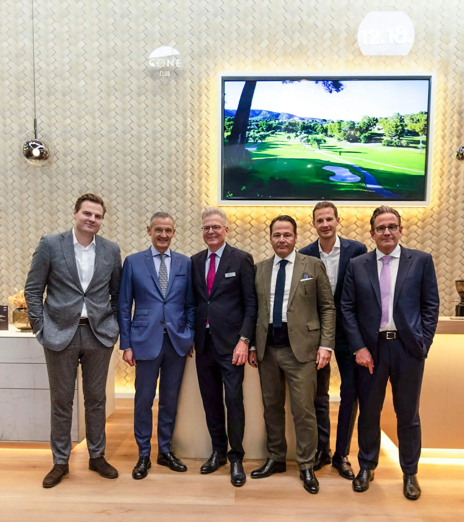 Sechs Männer in Anzügen stehen auf einer Messe vor einem Bildschirm mit einem Golfplatz.