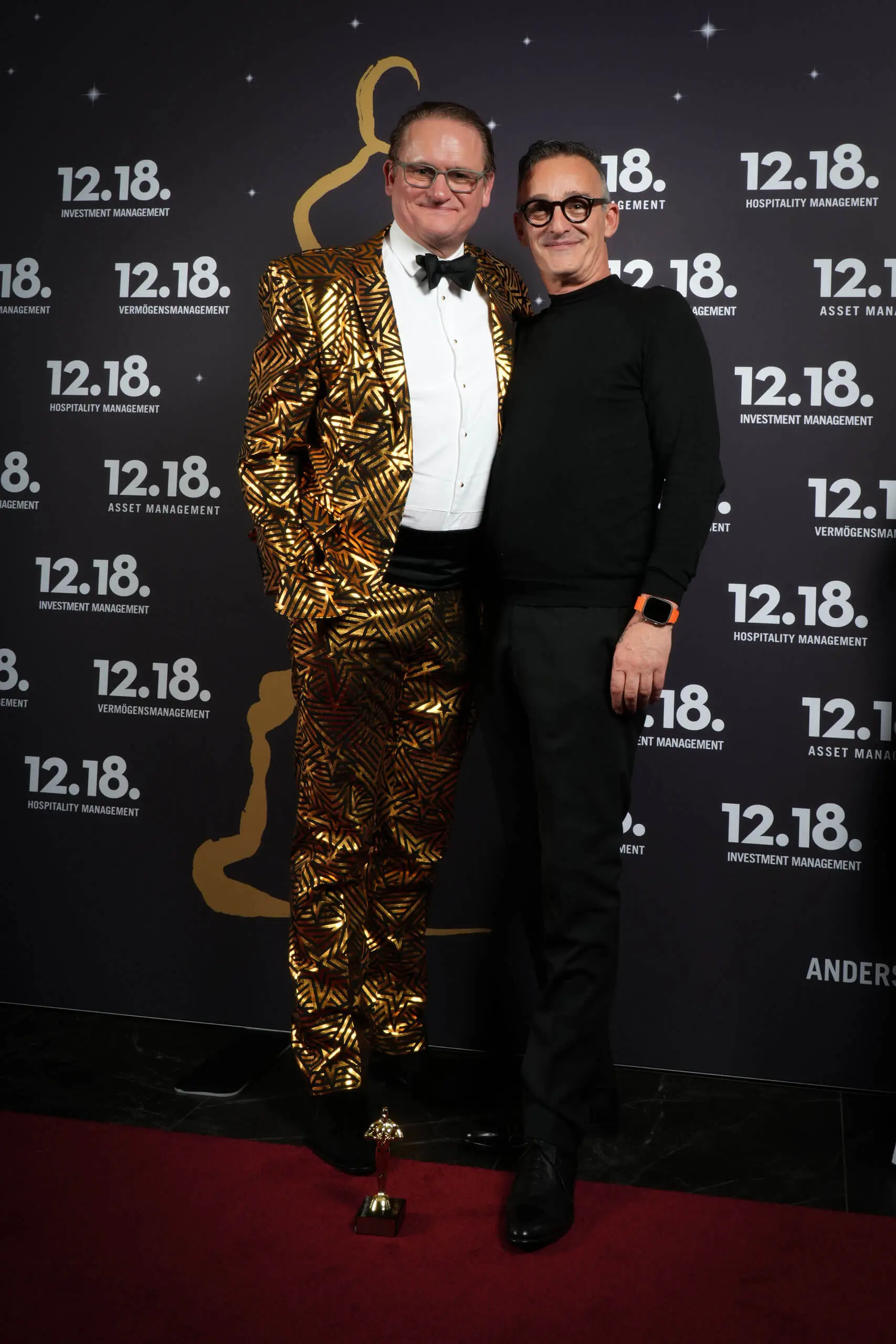 Zwei Männer in stilvoller Kleidung vor einem Event-Hintergrund mit 12.18-Branding.