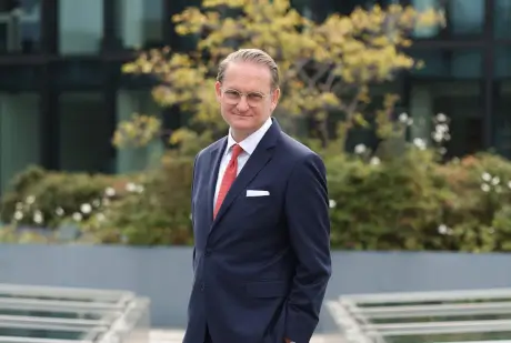 Man in a formal suit outdoors, standing confidently in front of green foliage.
