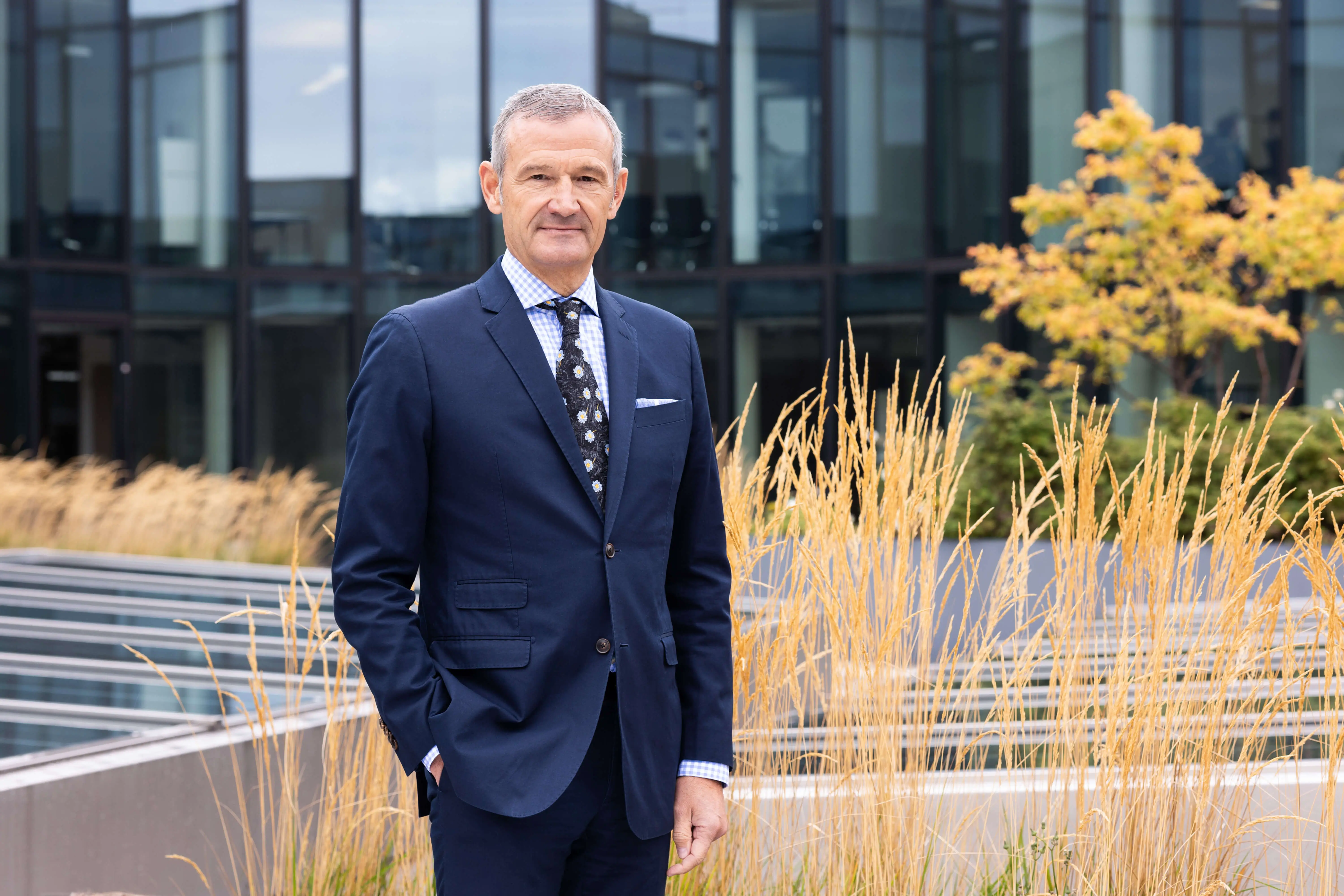 A professional man stands in front of a modern building, surrounded by golden grass.