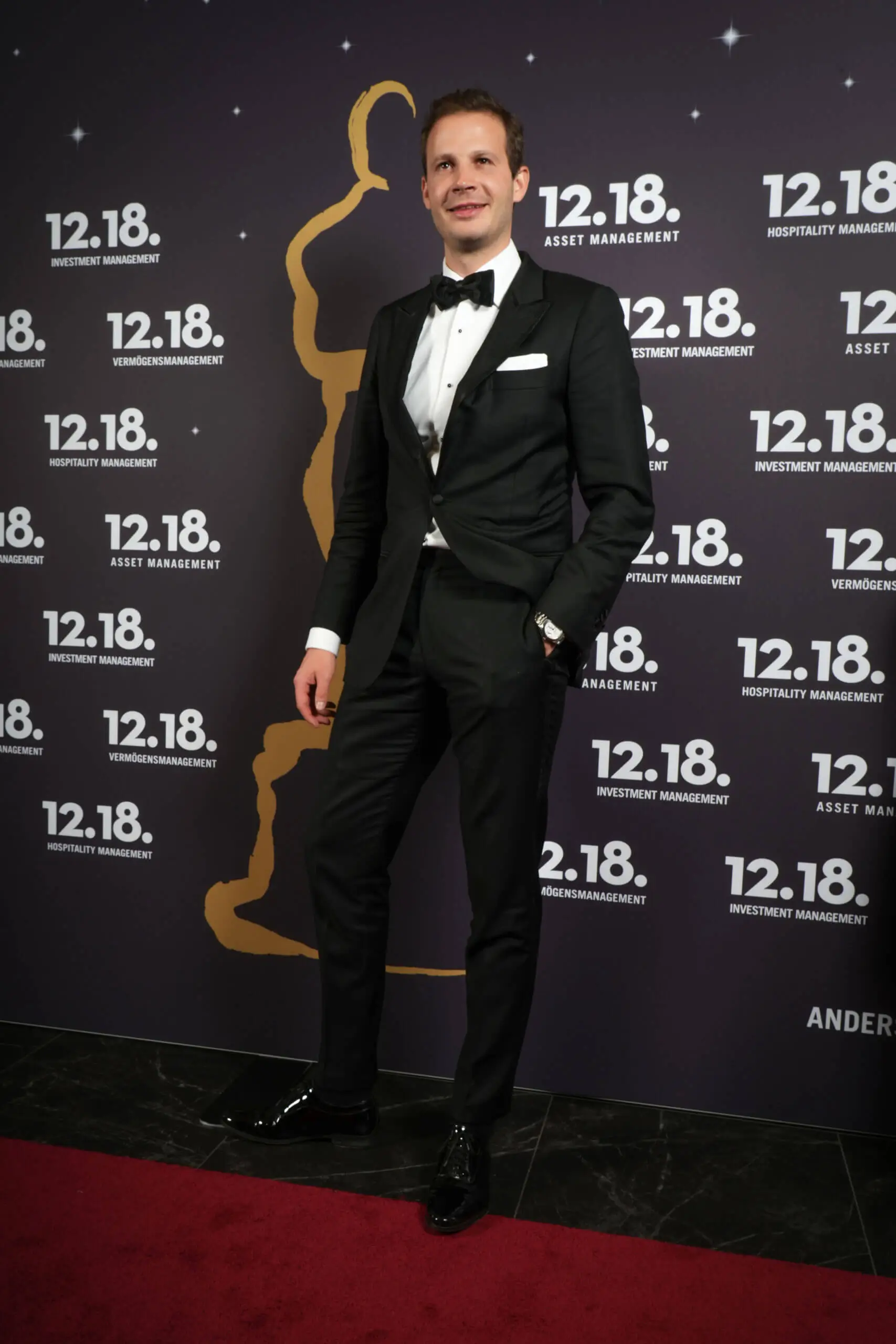 Ein Mann im Smoking posiert selbstbewusst vor einem eleganten Veranstaltungs-Hintergrund.