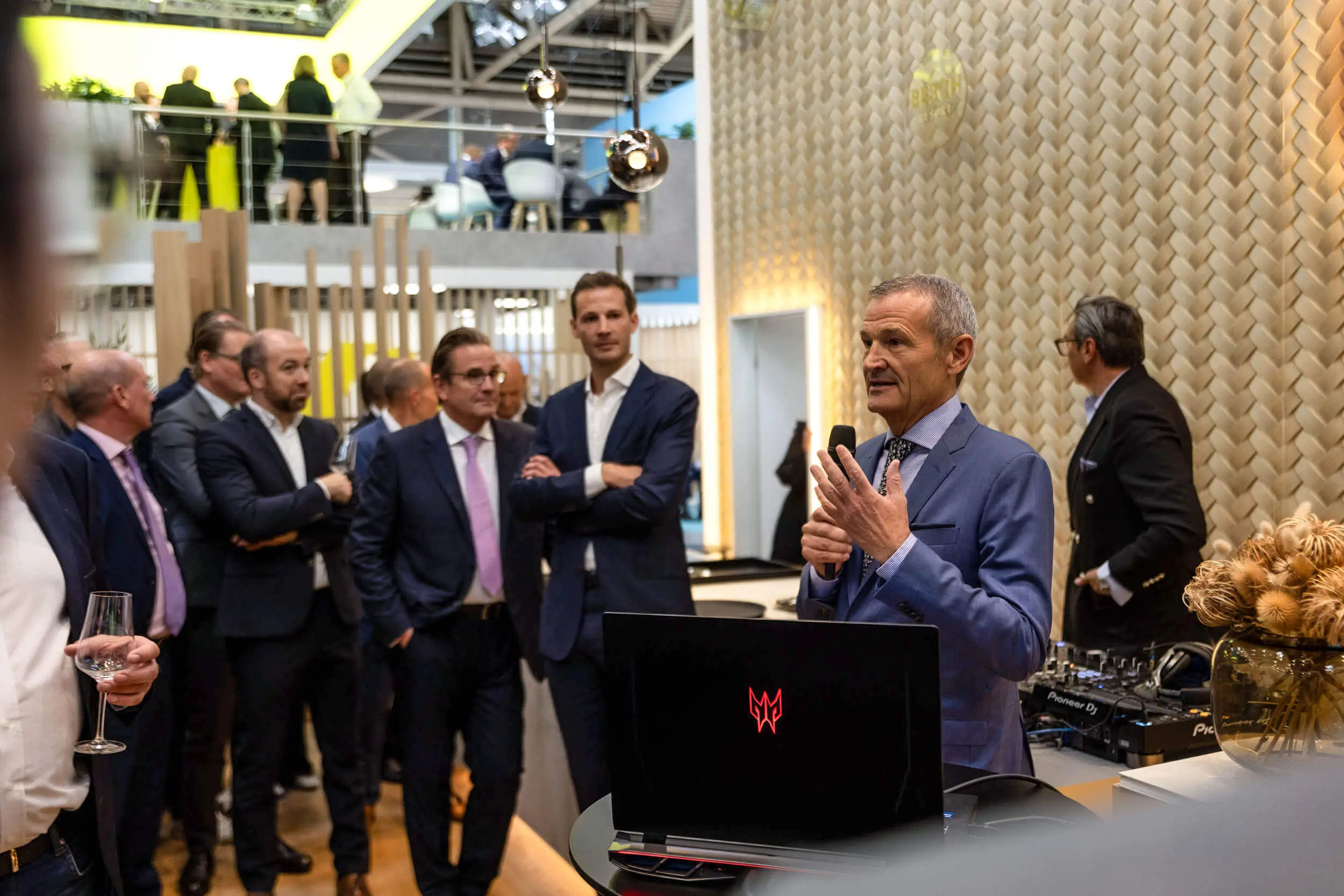 Ein Redner im blauen Anzug spricht an einem Messestand zu Gästen.
