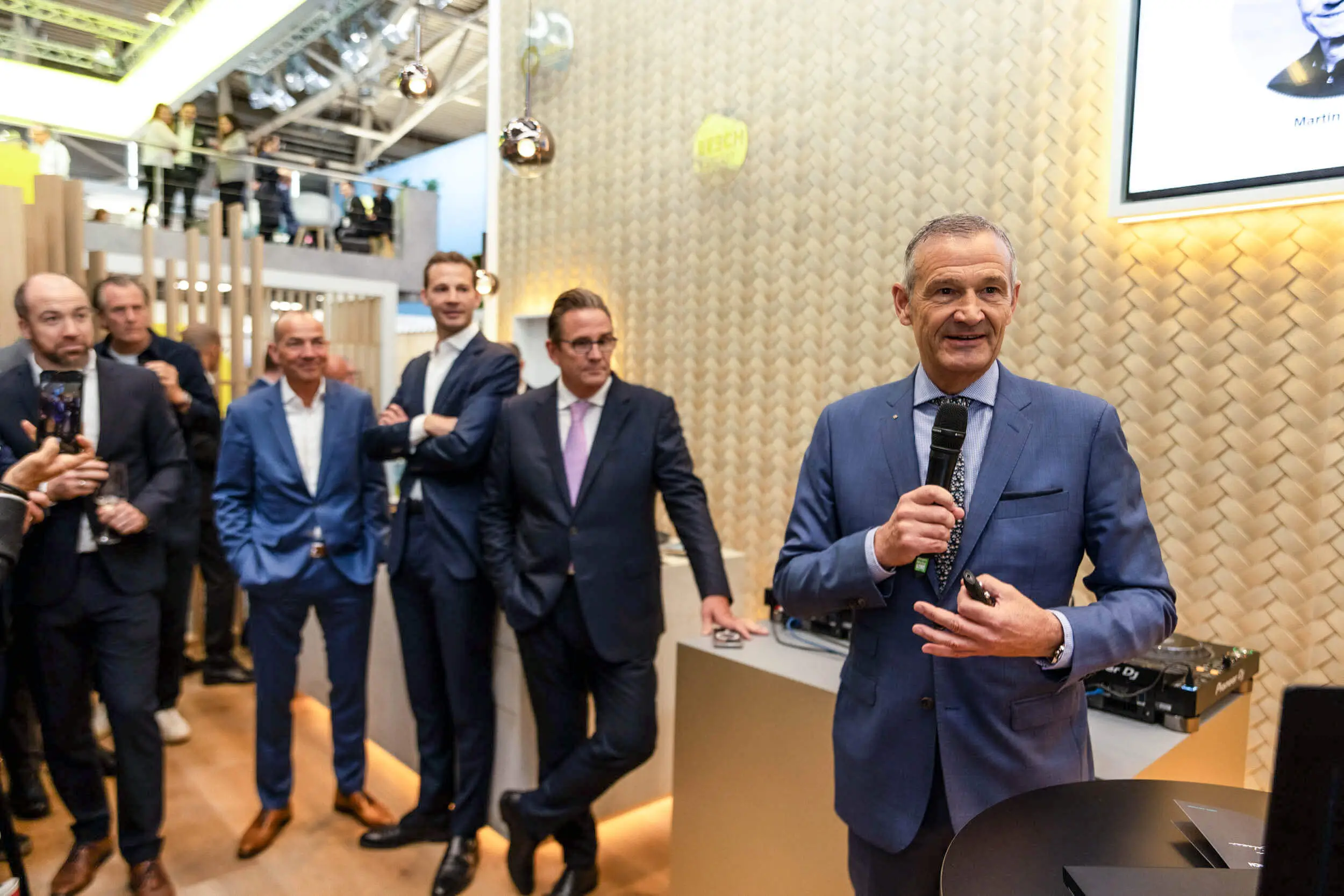 Ein Mann im blauen Anzug spricht auf einer Messe ins Mikrofon, während mehrere Besucher im Hintergrund zuschauen.