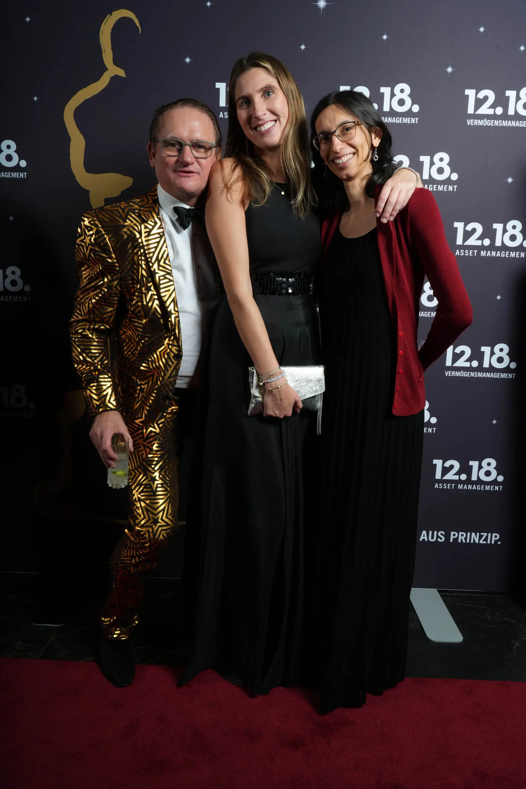 Drei stilvolle Teilnehmer bei einer glamourösen Veranstaltung mit einem goldenen Anzug und eleganten schwarzen Kleidern.