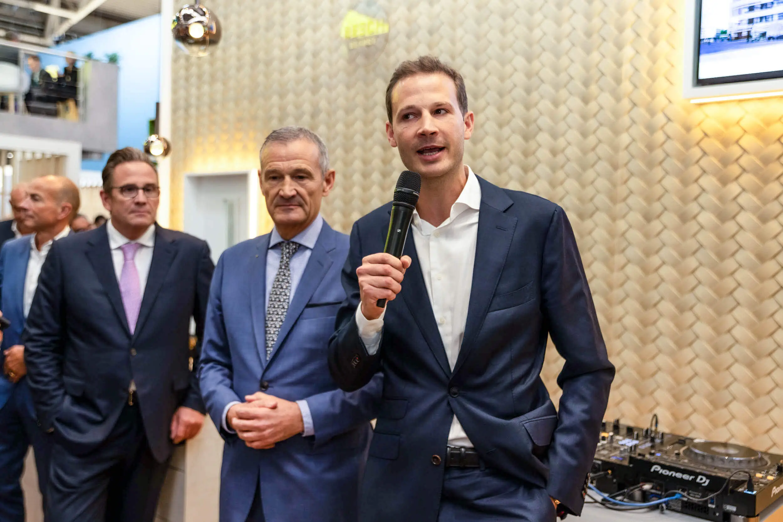 Ein Mann im Anzug spricht mit einem Mikrofon auf einer Messe, während Kollegen hinter ihm stehen.
