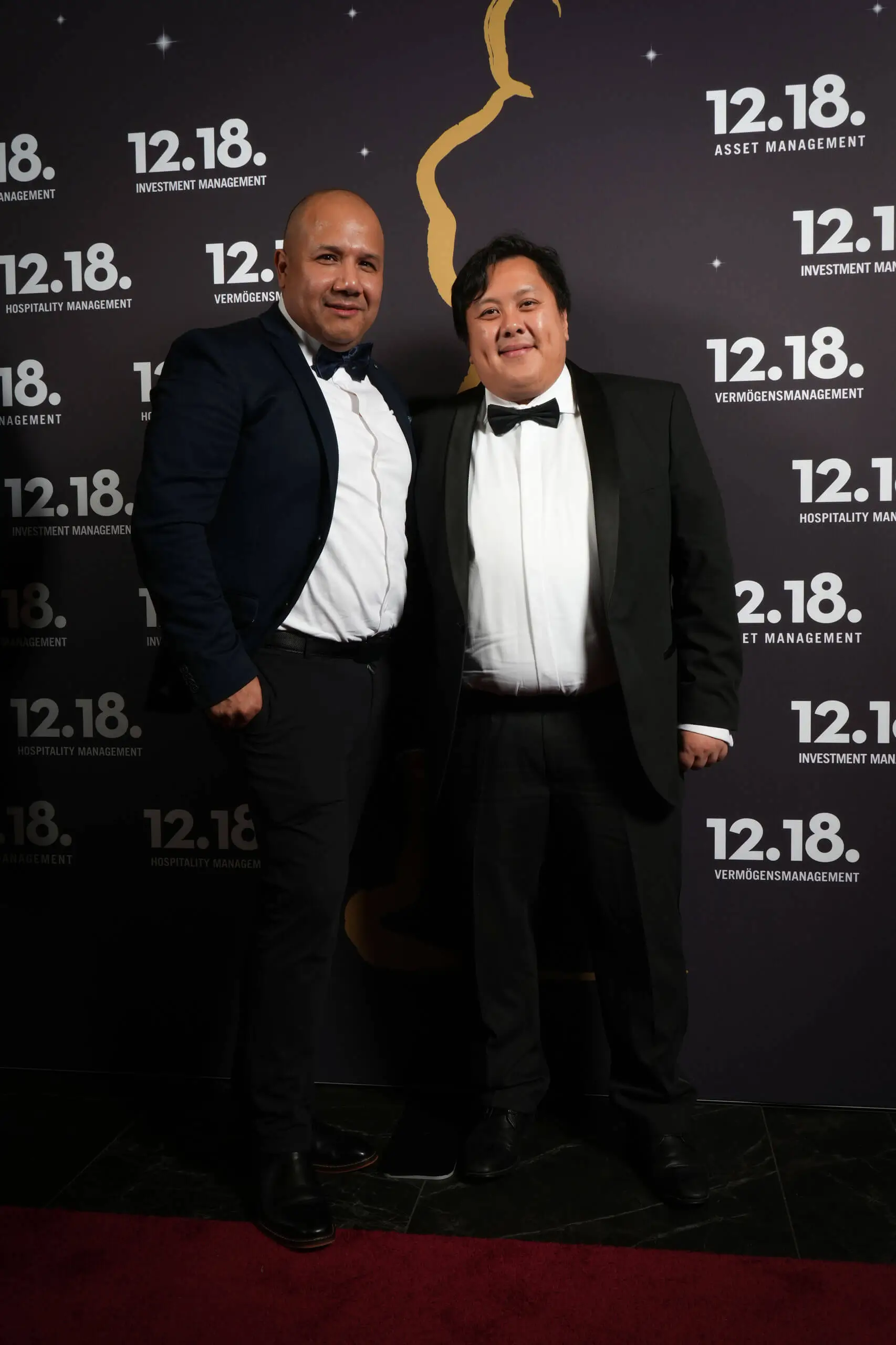 Zwei Herren in Smoking posieren elegant vor einem Veranstaltungshintergrund.